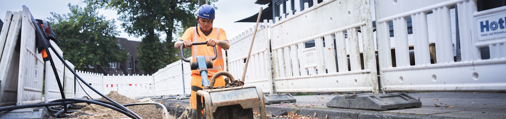 Person mit orangefarbener Warnweste bedient Baumaschine auf einer Baustelle neben einem weißen Zaun.