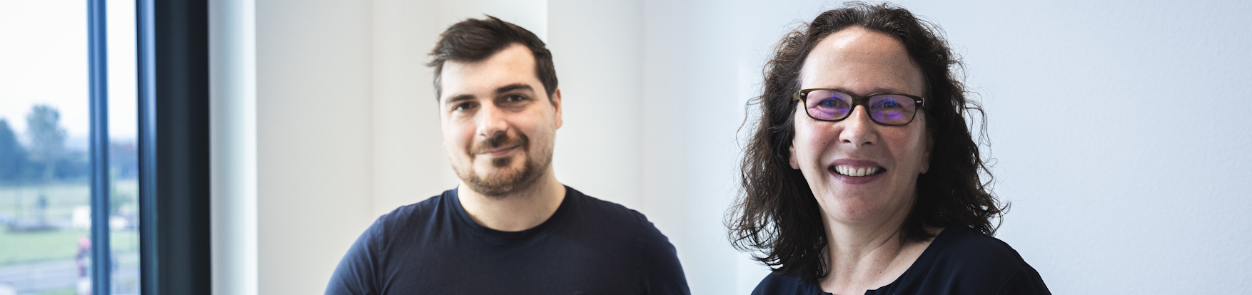 Zwei freundliche Personen stehen nebeneinander vor einem Fenster, moderne Büroatmosphäre