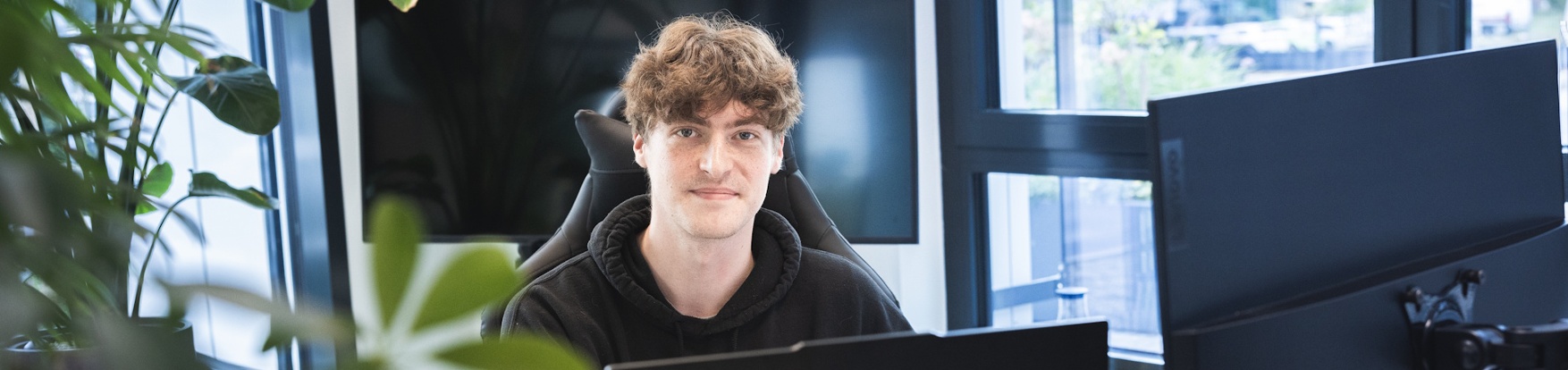Junger Mann sitzt an einem Schreibtisch mit mehreren Monitoren in einem modernen Büro mit großen Fenstern.